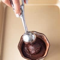 A hand holding an ice cream scoop of the truffle mixture and scooping in in the center of the cake batter in the ramekin before baking