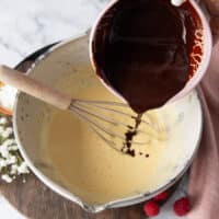 A hand pouring in the melted chocolate and butter mixture over the whipped egg and sugar mixture in the large cake batter bowl,
