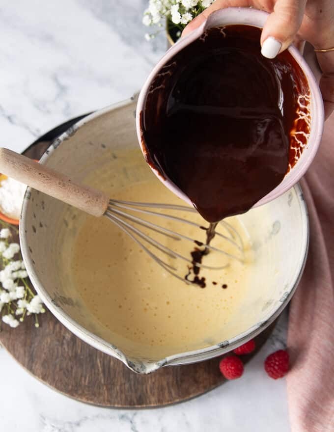 A hand pouring in the melted chocolate and butter mixture over the whipped egg and sugar mixture in the large cake batter bowl,