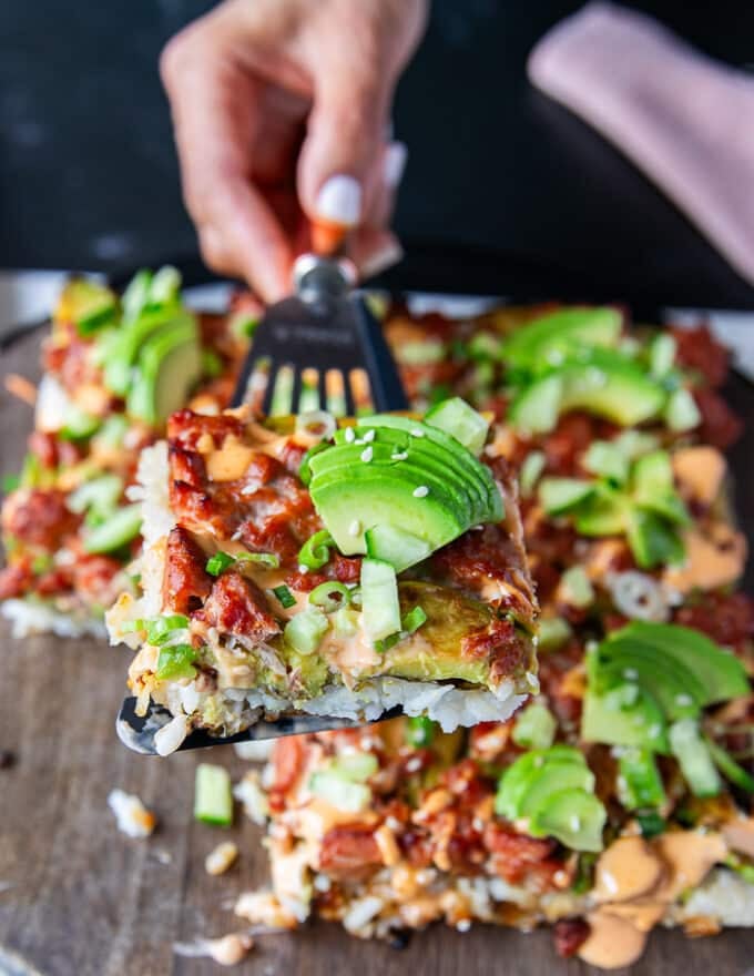 A hand holding a slice of sushi bake over a wooden tray using a spatula 