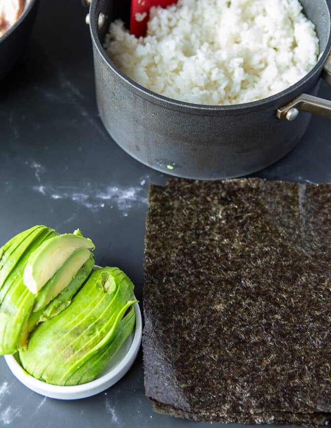 A pot of cooked sushi rice, some sliced avocados on a cooking board and some seaweed sheets 