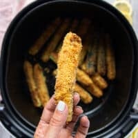 A hand holding one zucchini fries right out of the air fryer showing the texture and color of it