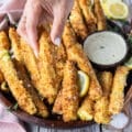A hand holding one crunchy zucchini on a plate of zucchini fries surrounded by parmesan dip