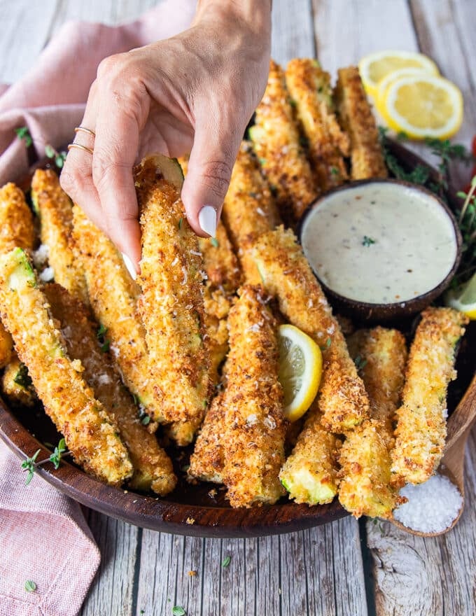 A hand holding one crunchy zucchini on a plate of zucchini fries surrounded by parmesan dip