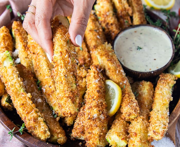 A hand holding one crunchy zucchini on a plate of zucchini fries surrounded by parmesan dip