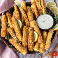 A plate of zucchini fries looking crisp, golden and perfect surrounded by lemon slices and a garlic parmesan dip
