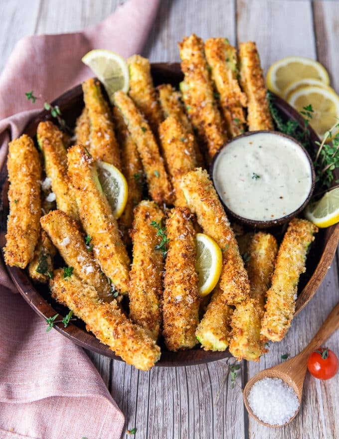 A plate of zucchini fries looking crisp, golden and perfect surrounded by lemon slices and a garlic parmesan dip 