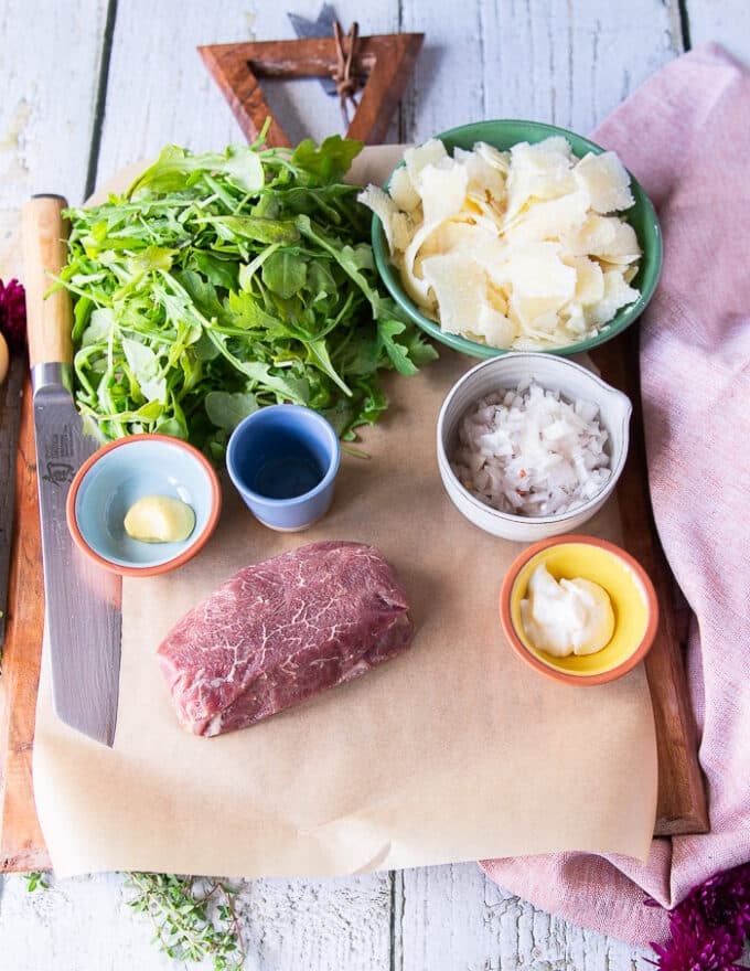 Beef carpaccio ingredients on a board including a piece of tenderloin, some arugula, some shaved parmesan in bowls, some mustard in a bowl, minced shallot in a bowl, white balsamic vinegar in a bowl, olive oil in a bottle, and salt and pepper