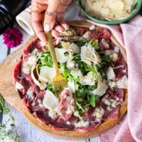 A hand holding a fork with a beef carpaccio close up showing the thinly sliced meat, the light vinaigrette and topping of diced onions, arugula and parmesan shavings