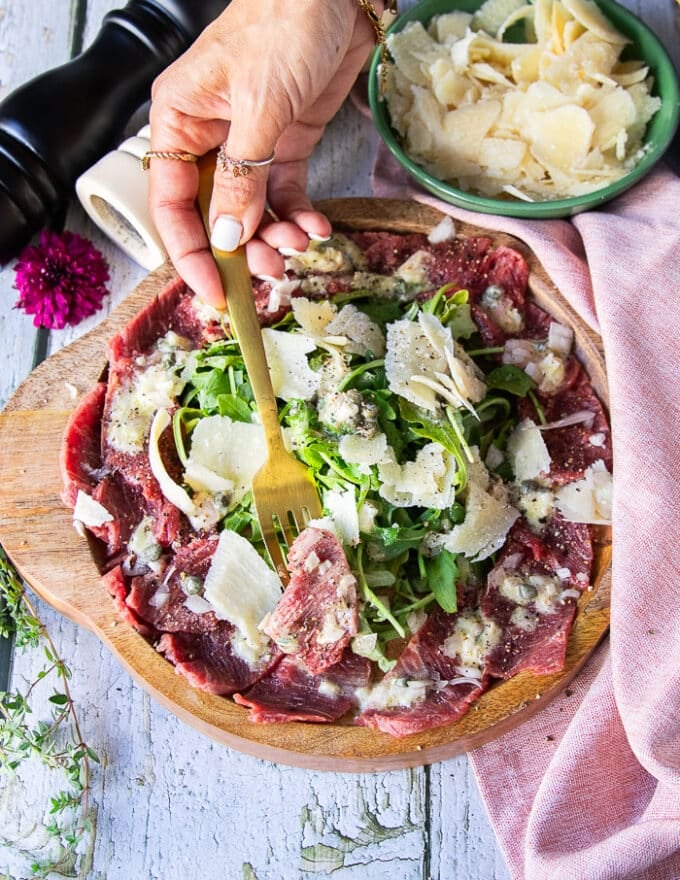 A hand holding a fork with a beef carpaccio close up showing the thinly sliced meat, the light vinaigrette and topping of diced onions, arugula and parmesan shavings