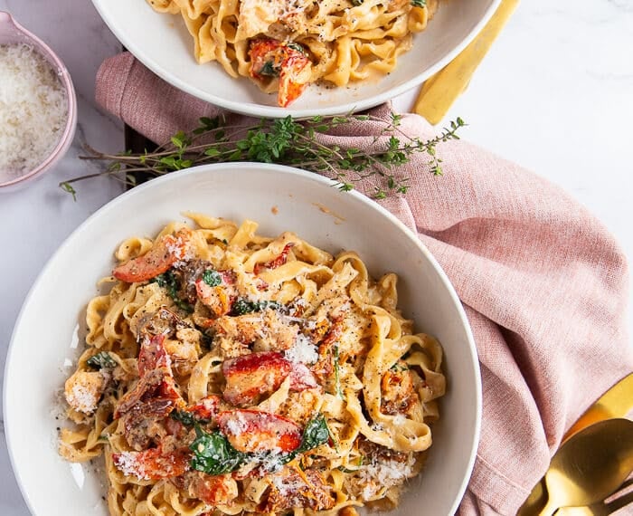two lobster pasta bowls garnished with parmesan cheese and surrounded by golden cutlery. The pasta is creamy and divine with cooked lobster meat on top