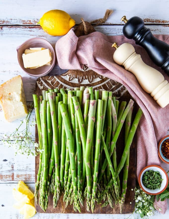 Ingredients for roasted asparagus including asparagus that's trimmed on a cutting board, some melted butter in a bowl. salt and pepper grinders, some fresh herbs in a bowl, some parmesan cheese block and chilli flakes in a small bowl 