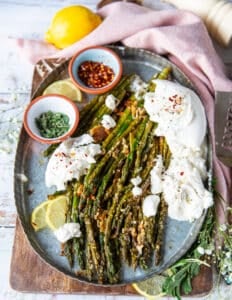 A plate of roasted asparagus with parmesan cheese served with some burrata cheese on the side, some extra chilli flakes for sprinkling, some herbs and parmesan cheese on top and lemon slices on the side