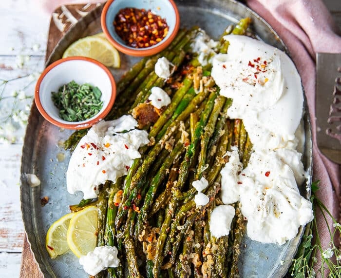 A plate of roasted asparagus with parmesan cheese served with some burrata cheese on the side, some extra chilli flakes for sprinkling, some herbs and parmesan cheese on top and lemon slices on the side