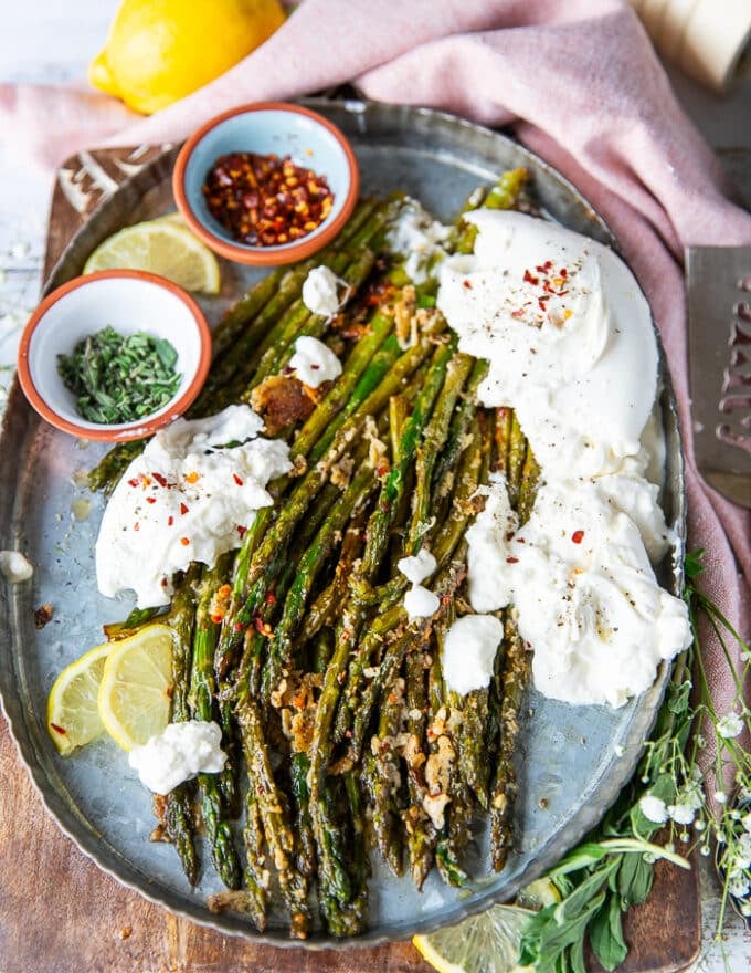 A plate of roasted asparagus with parmesan cheese served with some burrata cheese on the side, some extra chilli flakes for sprinkling, some herbs and parmesan cheese on top and lemon slices on the side