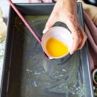 A hand pouring some melted butter at the bottom of a baking pan
