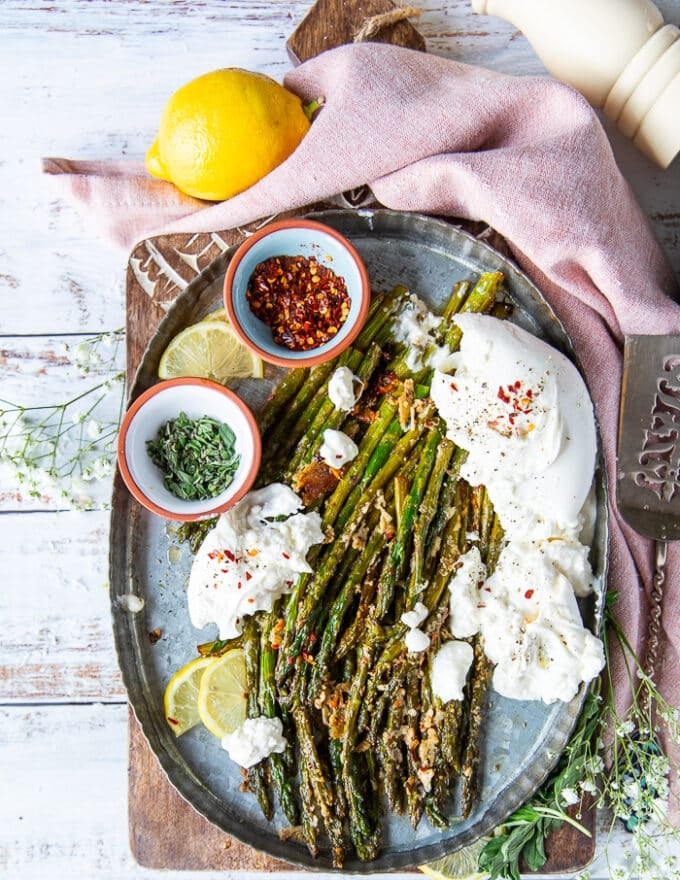 Top view of a plate of roasted asparagus served with a side dish of burrata cheese and lemon slices. Some chilli flakes on the side and fresh herbs