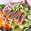 close up of a sliced ahi tuna salad showing the pink inside of the ahi tuna and the sesame crust on the outside, drizzled with a goma sesame dressing on a bed of avocado, cucumber, carrots, crispy wontons and more