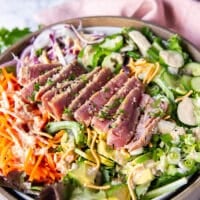 close up of a sliced ahi tuna salad showing the pink inside of the ahi tuna and the sesame crust on the outside, drizzled with a goma sesame dressing on a bed of avocado, cucumber, carrots, crispy wontons and more