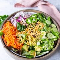 Salad ingredients including some cucumbers, some shredded carrots, greens, green onions, crispy wontons, red onions, avocados, cilantro, jalapenos