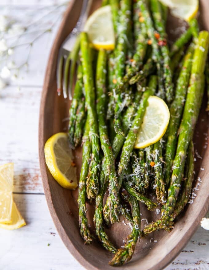 Close up of air fryer asparagus on a wooden plate with some parmesan sprinkled on top , chilli flakes and lemon slices showing the golden crisp tips of the asparagus
