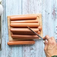 A hand holding a sharp knife and scorching the hot dogs on a diagonal to make like spirals on the side