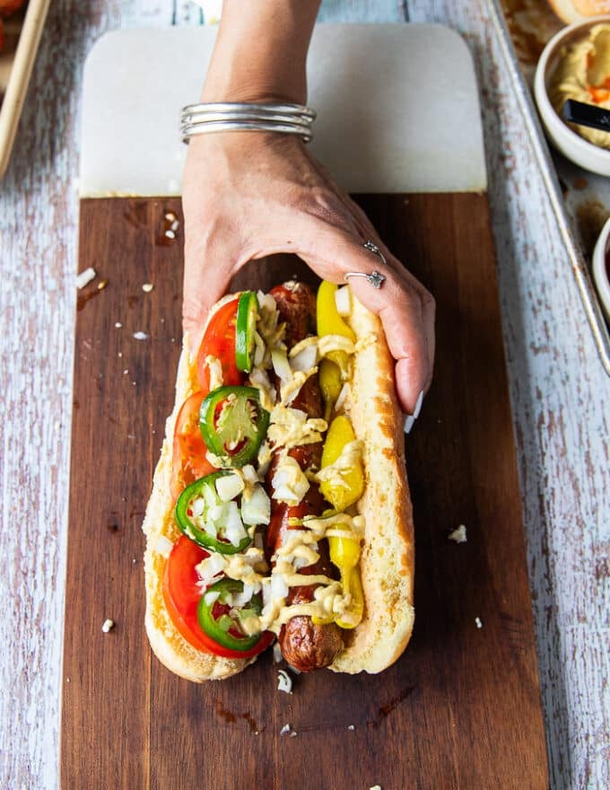 An assembled Chicago style hot dog showing the grilled hot dog with pickled peppers, sliced tomatoes, spicy mayo, jalapenos, minced onions