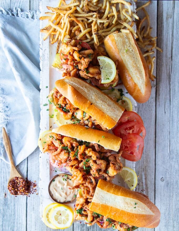 A large board with a few Po Boy Sandwiches cut up in half served along some french fries and a bowl of remoulade sauce for dipping 
