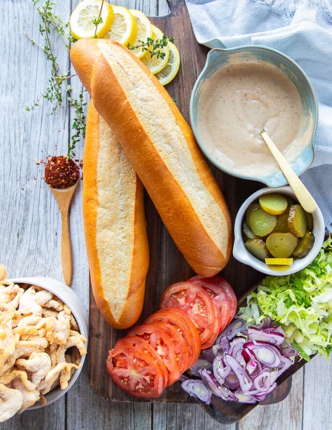 Ingredients to make a po boy sandwich including the foot long bread, the spicy remoulade sauce, some sliced pickles, some sliced tomatoes, lettuce and onions, and the battered shrimp on the side 
