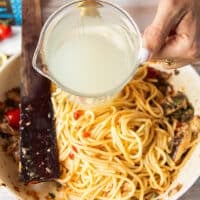 A hand pouring in some pasta water to the sauce to create a saucy sardine pasta