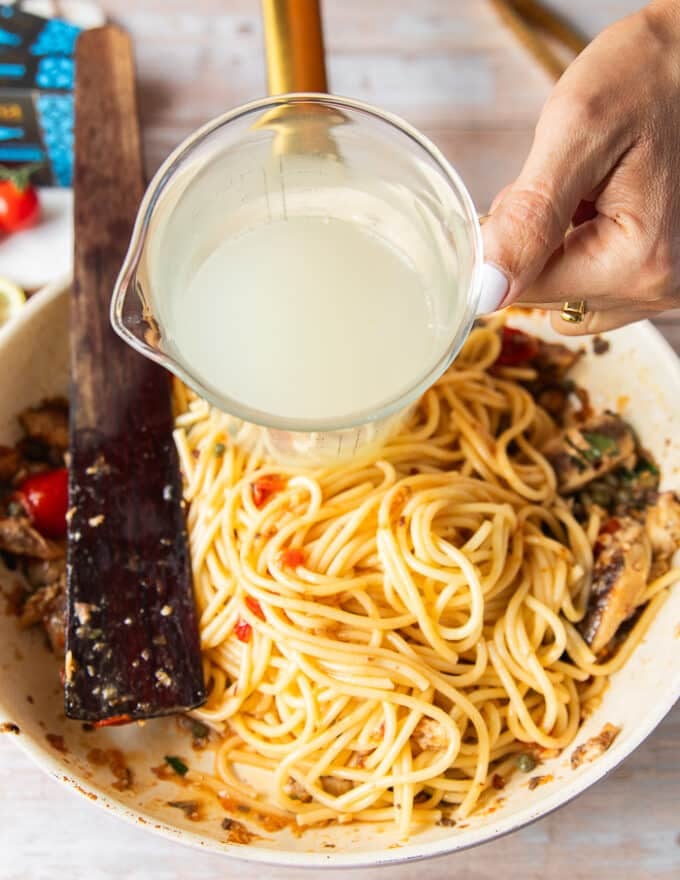 A hand pouring in some pasta water to the sauce to create a saucy sardine pasta