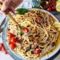 A hand holding a fork on a plate of sardine pasta showing the sardines, basil, tomatoes, capers, lemon and olive oil sauce around it