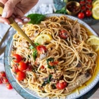 A hand holding a fork on a plate of sardine pasta showing the sardines, basil, tomatoes, capers, lemon and olive oil sauce around it
