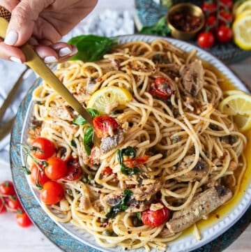 A hand holding a fork on a plate of sardine pasta showing the sardines, basil, tomatoes, capers, lemon and olive oil sauce around it