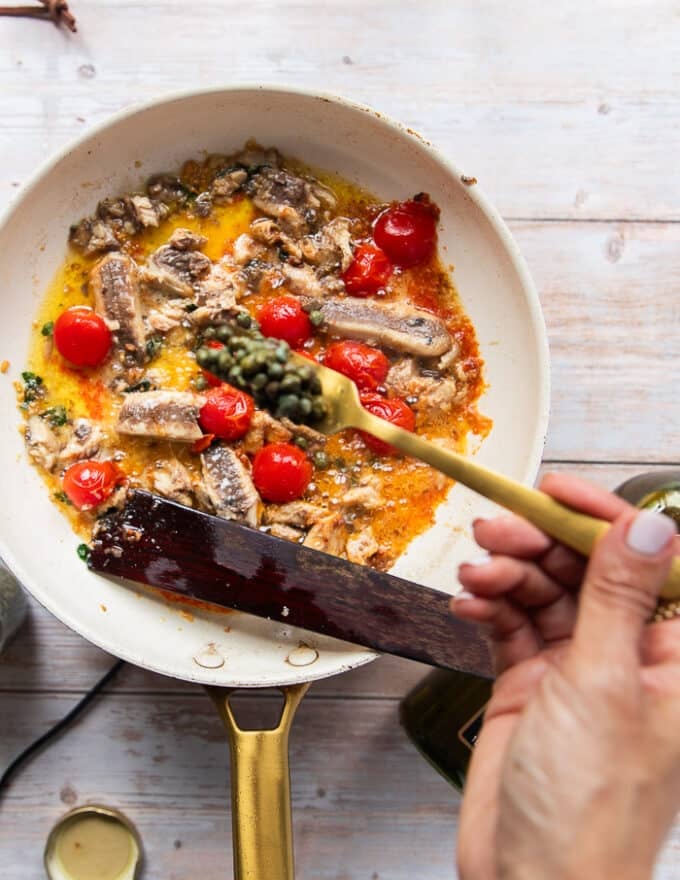 A hand adding in a spoonful of capers to the sauce pan