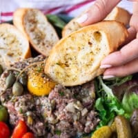 A hand holding a piece of crunchy baguette and pressing it on the eg yolk on stop of the steak tartare to break through and eat