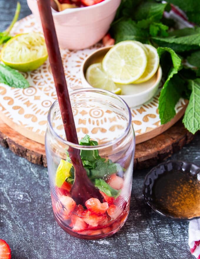 A wooden spoon muddling in the strawberries, mint and lime at the bottom of the cup to make the mojito recipe 