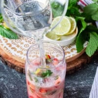 Club soda is poured over the muddled fruit and mint showing some fizz in the cup and continuing to pour until it fills up the cup.