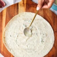 A hand spreading the sauce over the surface of the flour tortilla