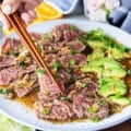 A hand holding two chopsticks and serving beef tataki sliced over a plate of ponzu sauce topped with crisp fried onions and green onions, surrounded by avocados sliced