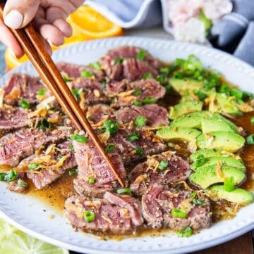 A hand holding two chopsticks and serving beef tataki sliced over a plate of ponzu sauce topped with crisp fried onions and green onions, surrounded by avocados sliced