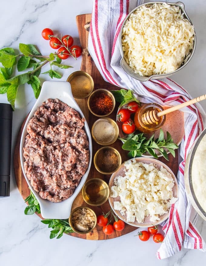 Ingredients for the grilled pizza or sausage pizza including ground sausage meat, sausage spices, feta cheese in a bowl, pizza doughy, fresh herbs, cherry tomatoes cut in half