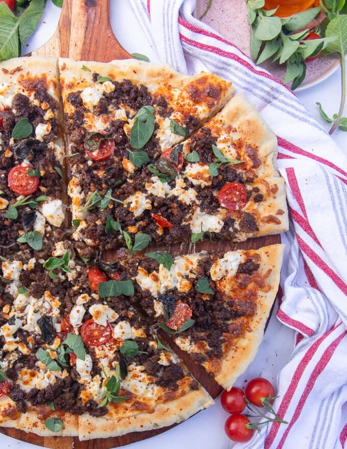 fully cooked grilled pizza on a wooden board surrounded by tea towel and some cherry tomatoes, a honey stick and some herbs 