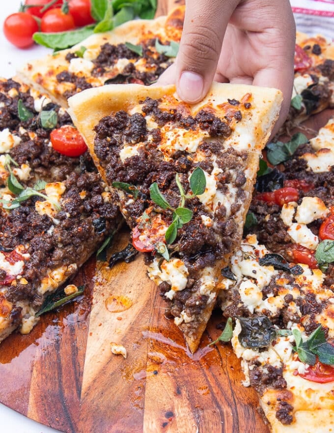 A hand holding a slice of grilled pizza close up showing the sausage on the pizza and the feta toppings