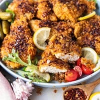 A plate with hot honey chicken tenders surrounded by lemon slices, a jar of hot honey and one chicken cut in half showing the juicy chicken inside.
