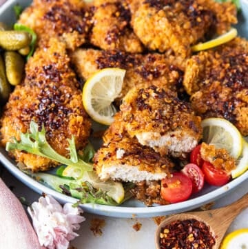 A plate with hot honey chicken tenders surrounded by lemon slices, a jar of hot honey and one chicken cut in half showing the juicy chicken inside.