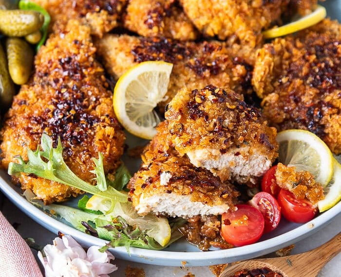 A plate with hot honey chicken tenders surrounded by lemon slices, a jar of hot honey and one chicken cut in half showing the juicy chicken inside.