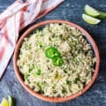 A plate of cilantro lime rice top view showing the fluffy cooked rice, the specks of green from cilantro, scallions, lime zest, jalapenos and surrounded by lime slices, jalapenos and a tea towel