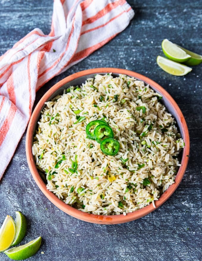 A plate of cilantro lime rice top view showing the fluffy cooked rice, the specks of green from cilantro, scallions, lime zest, jalapenos and surrounded by lime slices, jalapenos and a tea towel 
