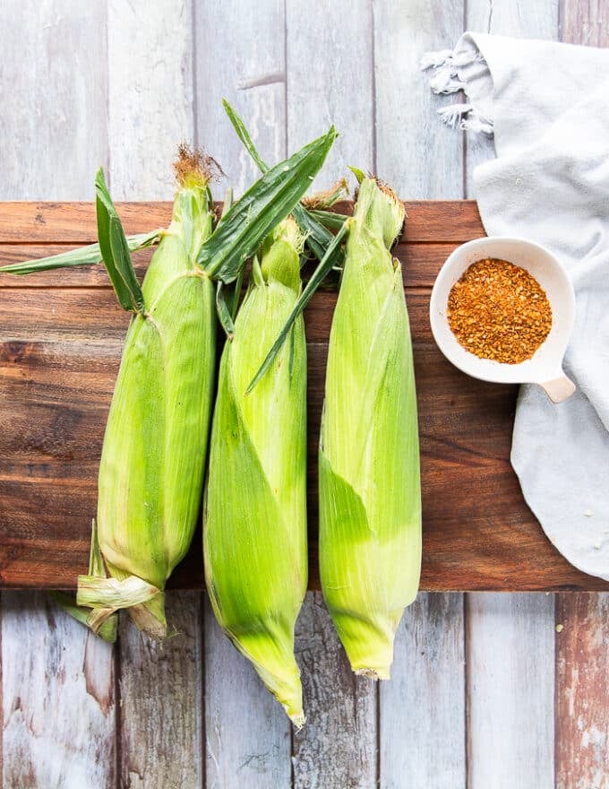Fresh corn on the cobs on a wooden board surrounded by a small plate of chilli powder and some olive oil 